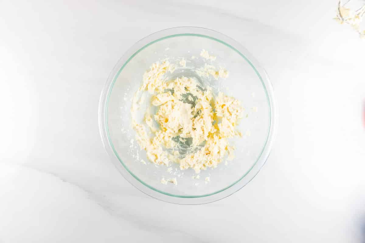 A glass bowl with partially mixed creamy butter and sugar sits on a white marble countertop, reminiscent of the beginnings of a delectable Strawberry Pretzel Salad Dip.