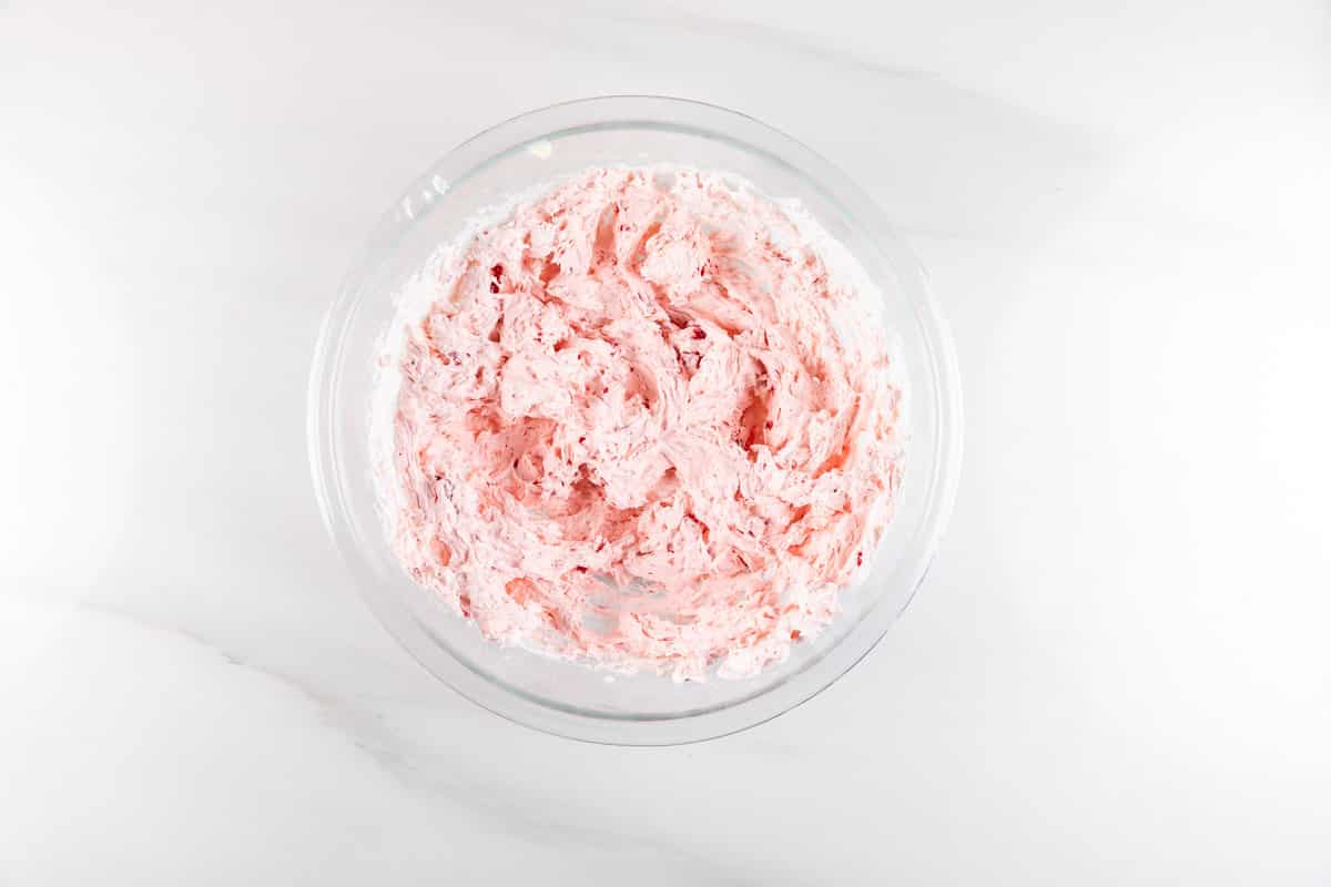 A glass bowl filled with light pink whipped frosting, reminiscent of strawberry pretzel salad dip, rests on a smooth, white marble surface. The frosting appears fluffy and creamy, with visible swirls and texture throughout.