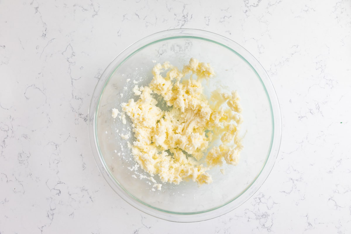 A glass bowl containing a light, fluffy mixture of creamed butter and sugar sits on a white marble countertop. The pale yellow mixture is smooth and perfect for creating delightful Chocolate Chip Snickerdoodles, adding an irresistible twist to your baking adventure.