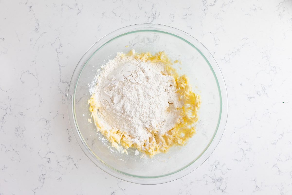 A glass bowl on a marble countertop contains a mixture of flour, butter, and hints of cinnamon and chocolate chips. The ingredients are partially combined, suggesting the beginning stages of crafting delicious Chocolate Chip Snickerdoodle dough.