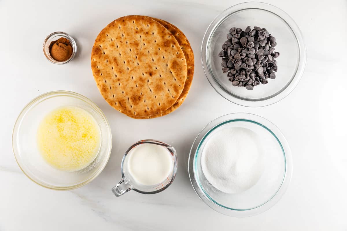 Ingredients for a dessert recipe arranged on a white surface: a bowl of chocolate chips, a stack of round biscuits resembling baked churro chips, a small bowl of cinnamon, a bowl of melted butter, a measuring cup of milk, and a bowl of sugar.