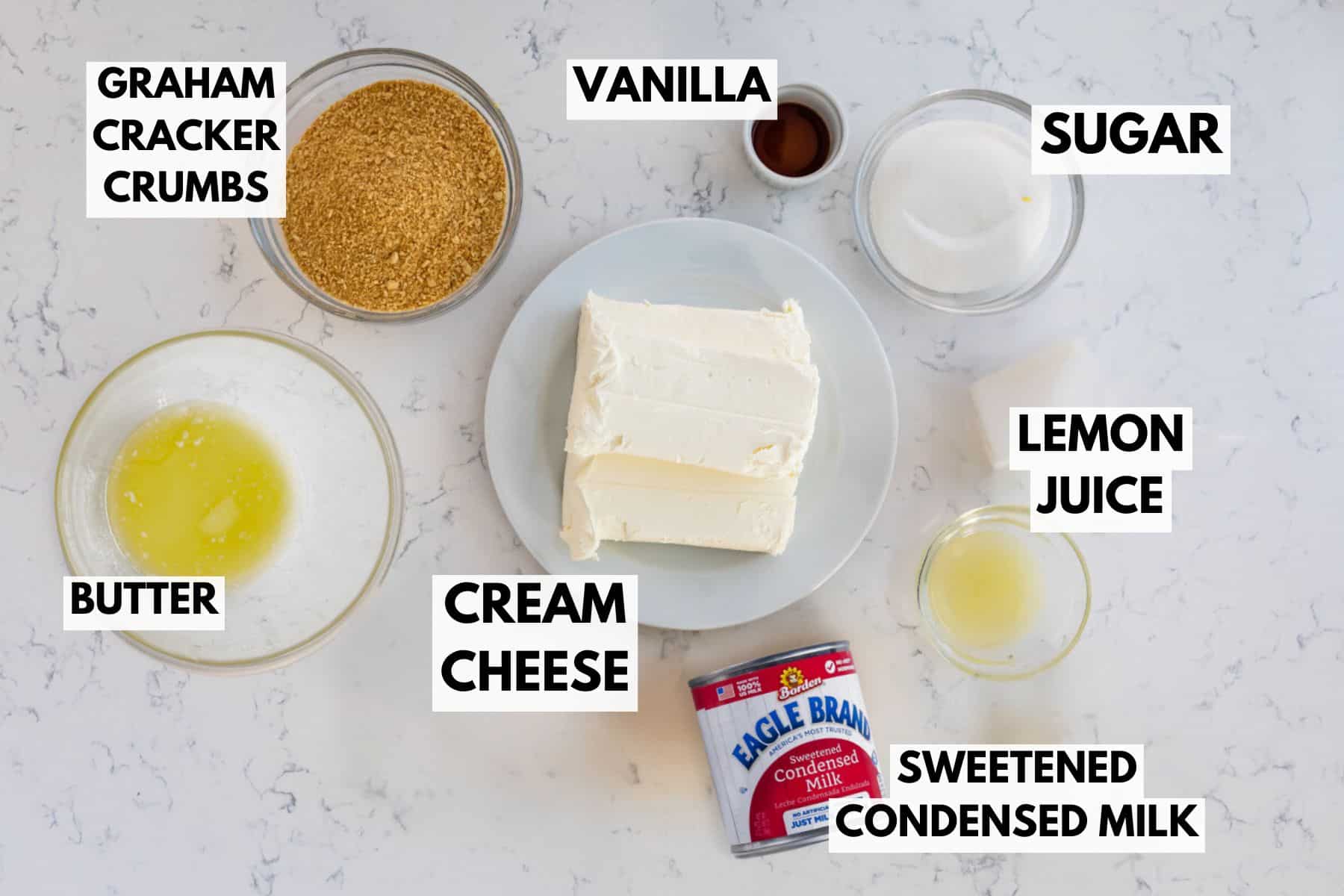 Top view of no bake cheesecake ingredients on a marble surface. Visible items include bowls of graham cracker crumbs, sugar, melted butter, lemon juice, and sweetened condensed milk, along with vanilla extract and blocks of cream cheese on a plate.