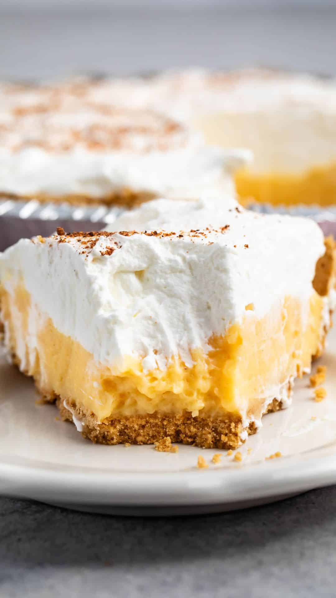 A close-up of a slice of creamy eggnog pie on a plate, featuring a graham cracker crust, a smooth yellow filling, and a generous layer of whipped cream on top, sprinkled with cocoa powder. Another pie is visible in the background.