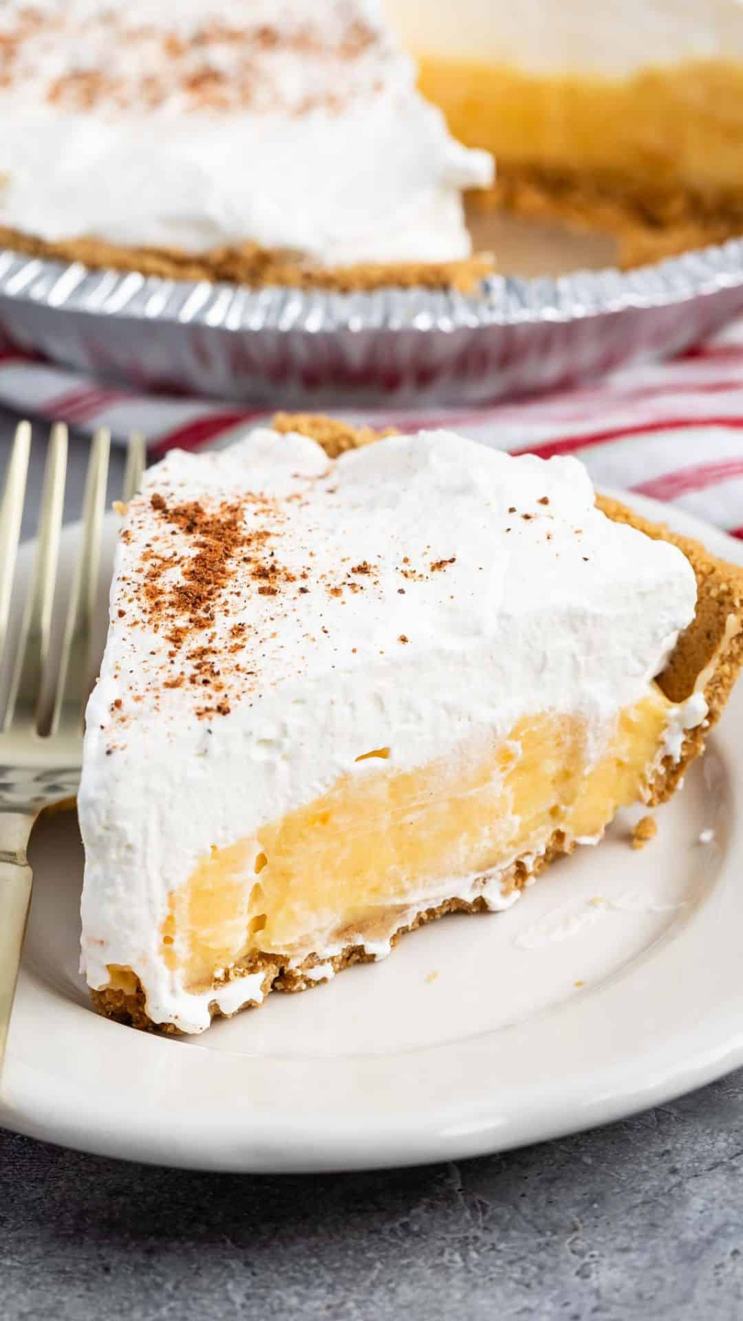 A slice of eggnog pie with a golden crust and fluffy whipped topping, lightly sprinkled with cocoa powder, is served on a white plate. The pie is set on a gray surface with a fork beside it, and the rest of the indulgent creation is visible in the background.