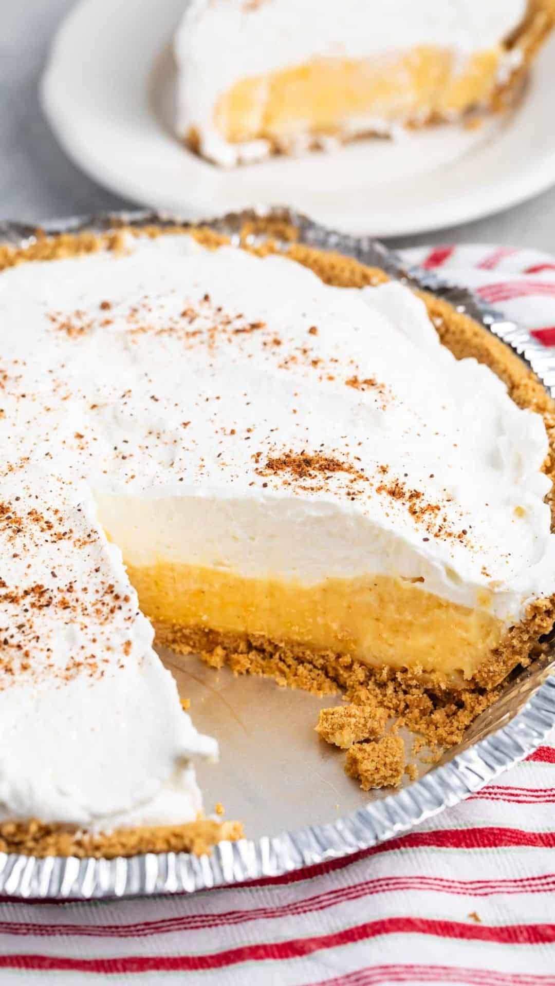 A freshly sliced eggnog pie in a silver pie dish features a golden-brown crust, a thick creamy filling, and is topped with a layer of fluffy whipped cream. A smaller slice sits on a white plate in the background, with a red-striped cloth nearby.