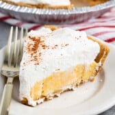 A slice of creamy Eggnog Pie crowned with whipped cream and a sprinkle of cocoa sits on a white plate. A fork rests beside it, tempting the senses. In the background, the remaining pie in its tin is partially visible, hinting at more indulgence to come.