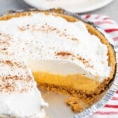 A creamy Eggnog Pie with a graham cracker crust and a sprinkle of cinnamon on top sits on a striped cloth. A slice is missing, revealing the rich yellow filling. In the background, theres a plate with another slice of this delicious pie.