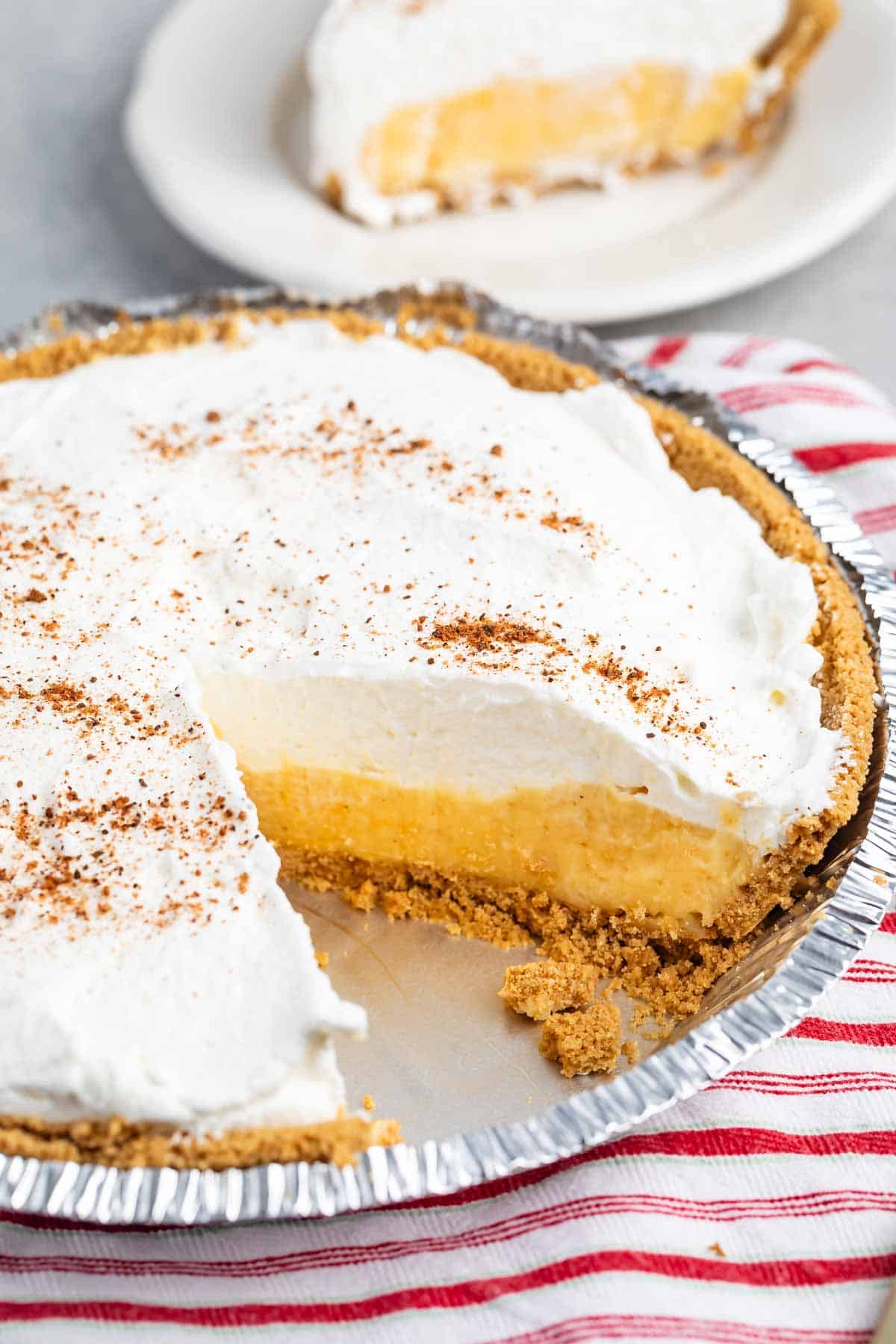 A creamy Eggnog Pie with a graham cracker crust and a sprinkle of cinnamon on top sits on a striped cloth. A slice is missing, revealing the rich yellow filling. In the background, theres a plate with another slice of this delicious pie.