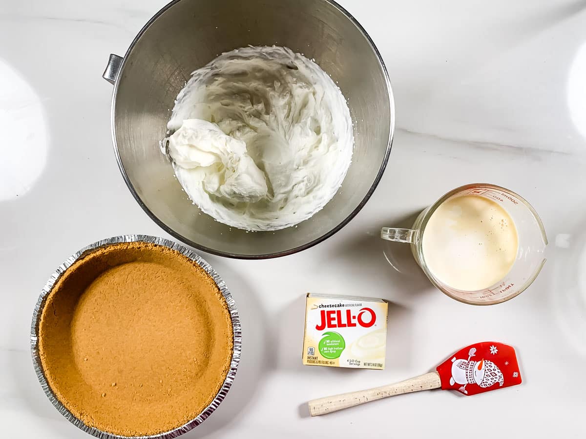 A cheesecake preparation setup on a white surface: a mixing bowl with whipped filling, an eggnog pie nestled in a graham cracker crust, a measuring cup with creamy liquid, a box of Jell-O cheesecake mix, and a spatula adorned with a snowman design.