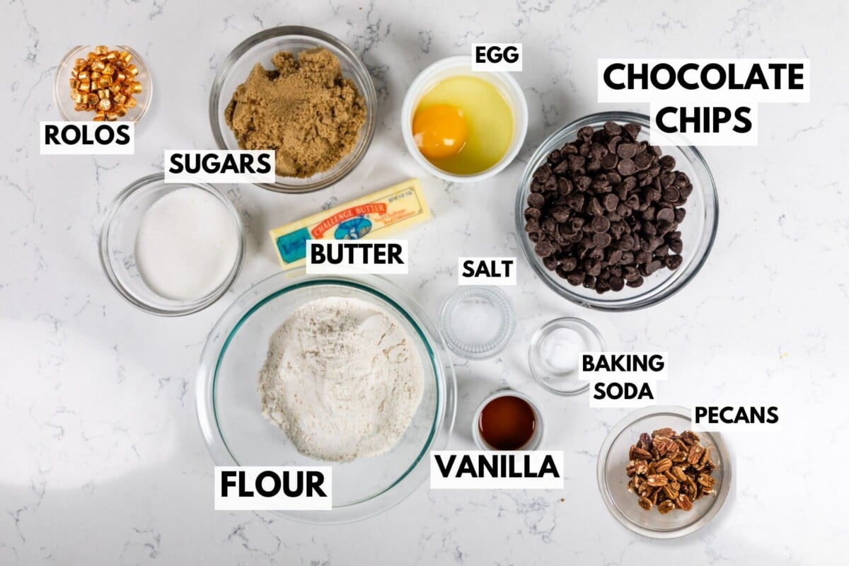 Turtle Cookies ingredients are neatly arranged on a marble counter, featuring labeled bowls of Rolos, brown and white sugars, butter, an egg, chocolate chips, flour, salt, vanilla extract, baking soda, and pecans.