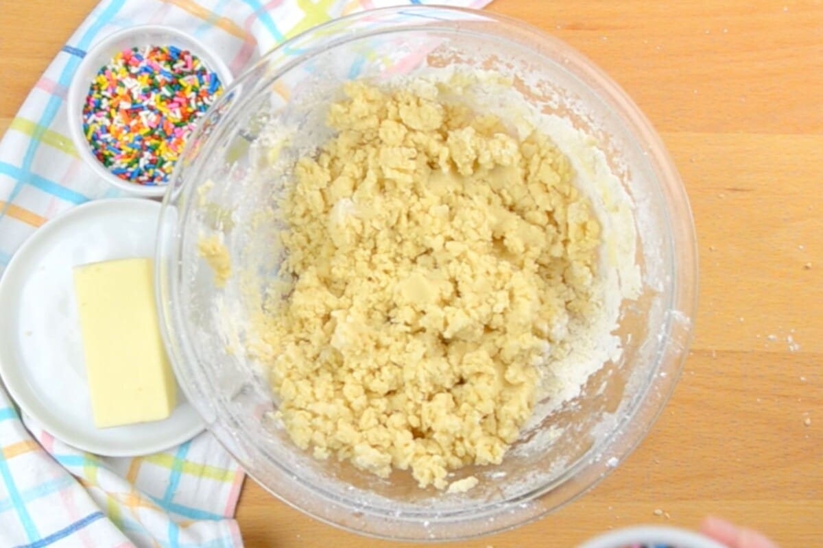 cookie dough in clear bowl on brown table with sprinkles next to it.