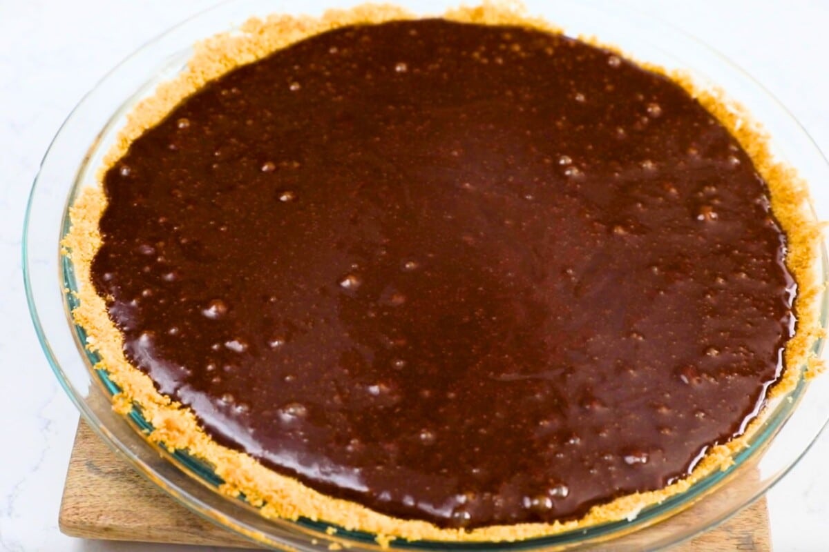 A glass pie dish filled with a rich, dark chocolate chess pie filling sits atop a wooden board. The pie boasts a golden-brown crumb crust along the edges. The chocolate surface appears smooth with a few visible bubbles.