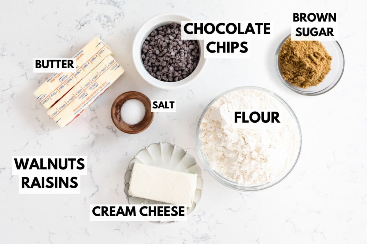 Ingredients for a delightful rugelach recipe on a white marble surface labeled: butter, chocolate chips, brown sugar, flour, walnuts, raisins, cream cheese, salt. They are displayed in bowls and wrappers.