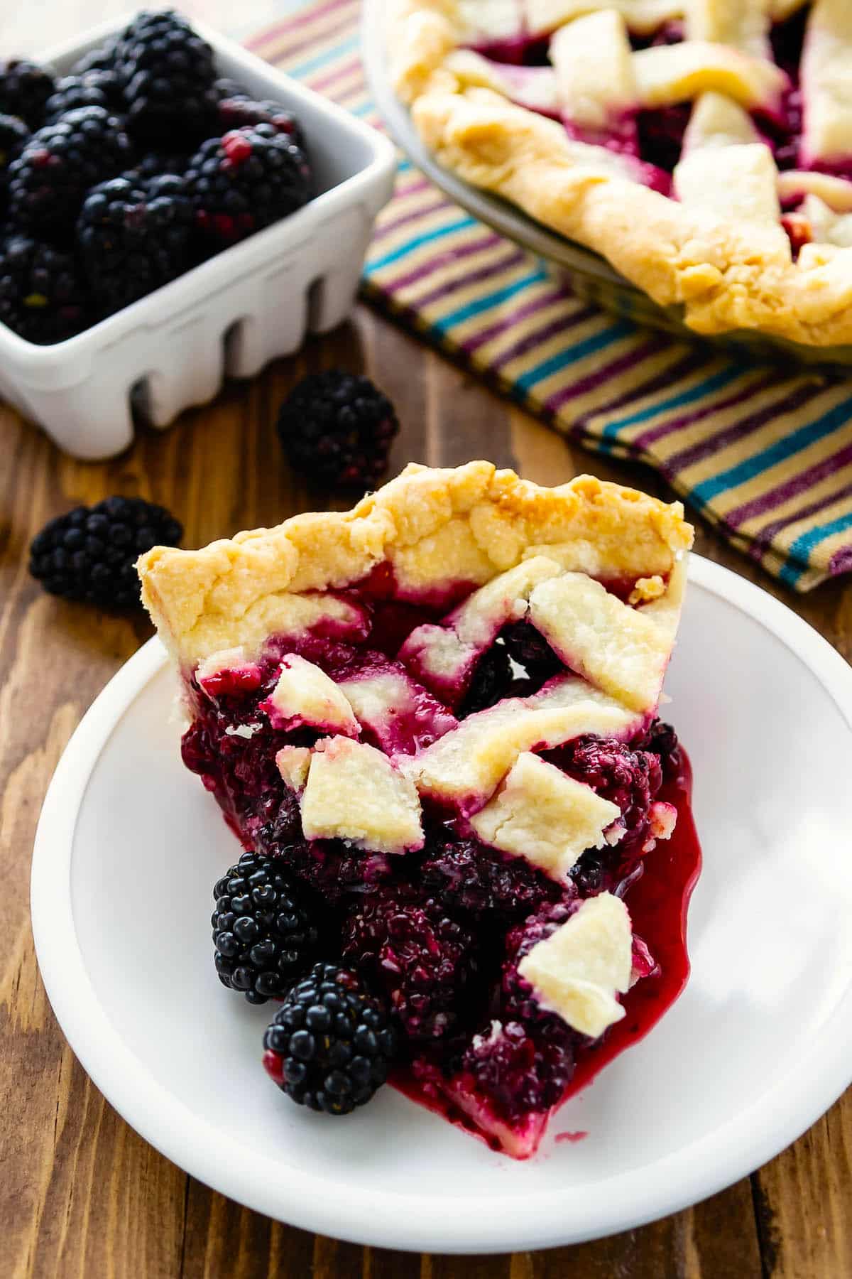 slice of blackberry pie with lattice crust on white plate on brown surface.