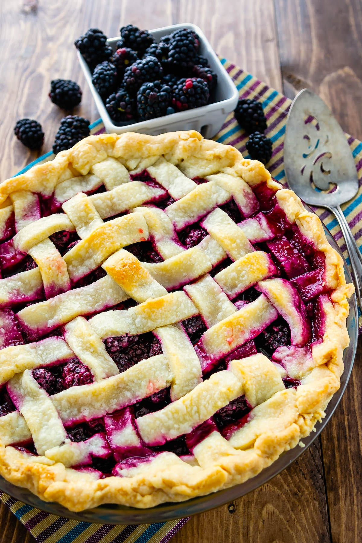 pie with lattice top with blackberries behind on wood surface