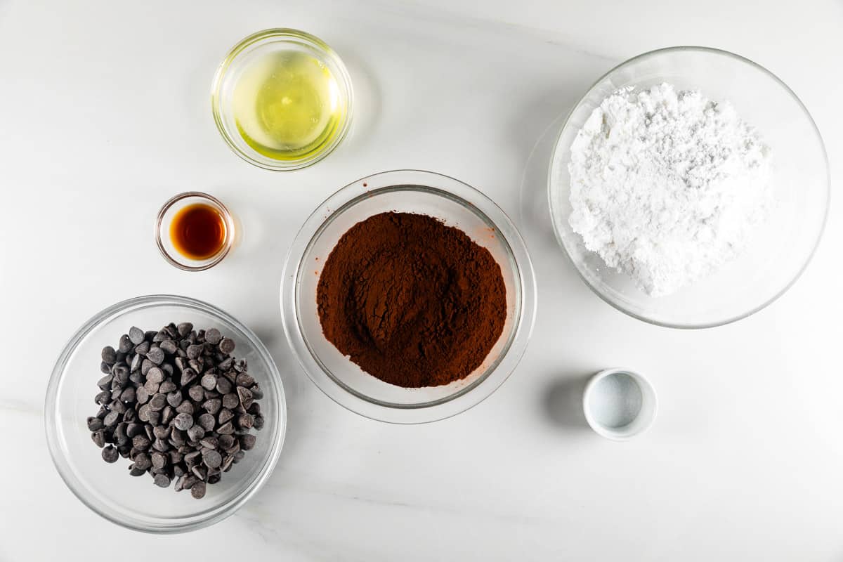 A top view of baking ingredients for flourless chocolate cookies on a white surface: a bowl of chocolate chips, a bowl of powdered sugar, cocoa powder, egg whites, vanilla extract, and salt—all ready to create decadent delights.