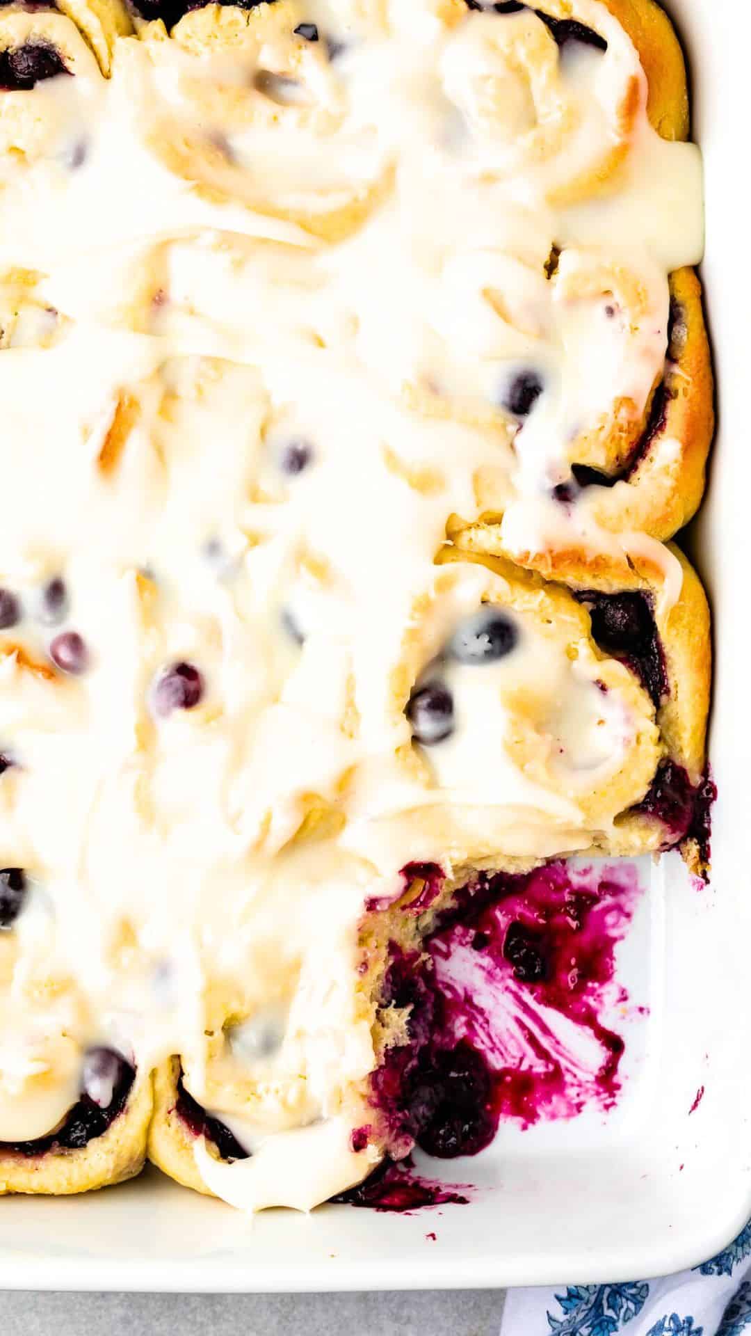 A close-up of Blueberry Sweet Rolls topped with creamy icing in a baking dish. One roll is missing, revealing a luscious purple fruity filling.