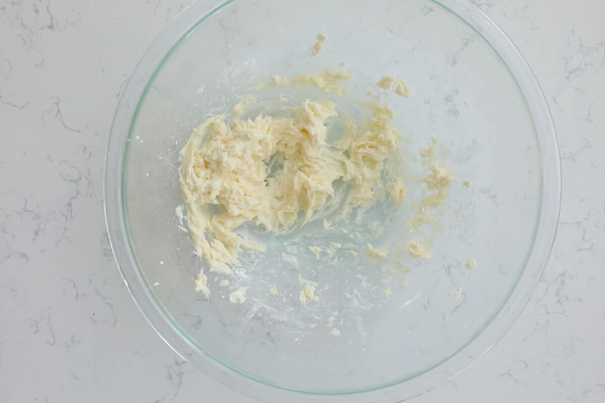 A clear glass bowl with partially mixed butter and sugar rests on a marbled countertop, ready to transform into a creamy cheesecake dip.