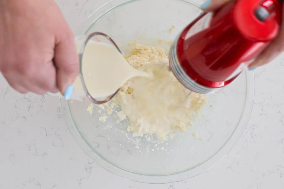 A person is pouring milk from a measuring cup into a glass bowl with flour, preparing to whip up a creamy cheesecake dip using a red hand mixer. The bowl rests on a white marble countertop.