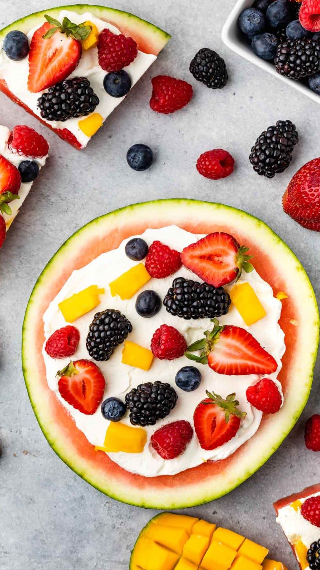 A vibrant watermelon pizza serves as the base, adorned with whipped cream, sliced strawberries, blackberries, blueberries, raspberries, and diced mango. Its surrounded by scattered berries and a sliced mango on a light gray surface.