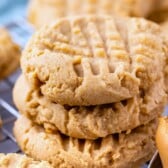 A stack of peanut butter cookies from a timeless recipe, featuring a crisscross fork pattern on top, sits on a wire cooling rack. The golden-brown treats bask in their fresh-baked glory against a blurred background with more cookies and a hint of blue.