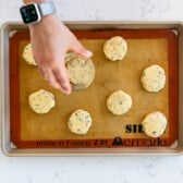 unbaked cookies on cookie sheet with hand pressing one flat with a glass