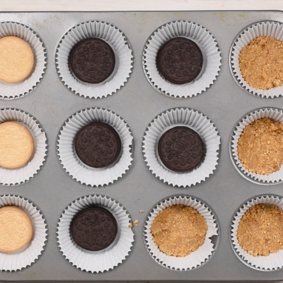 muffin pan with cupcake liners filled with graham cracker crust and Oreos.