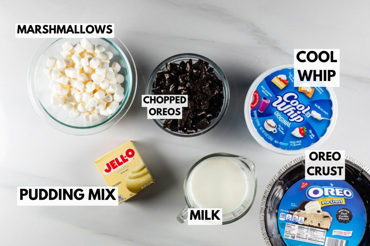 Top-down view of dessert ingredients on a white counter: bowls of mini marshmallows and chopped Oreos, a package of vanilla pudding mix, a carton of milk, and a tub of Cool Whip. Everything is set to create the perfect Oreo Pie with its crumbly crust labeled and ready.