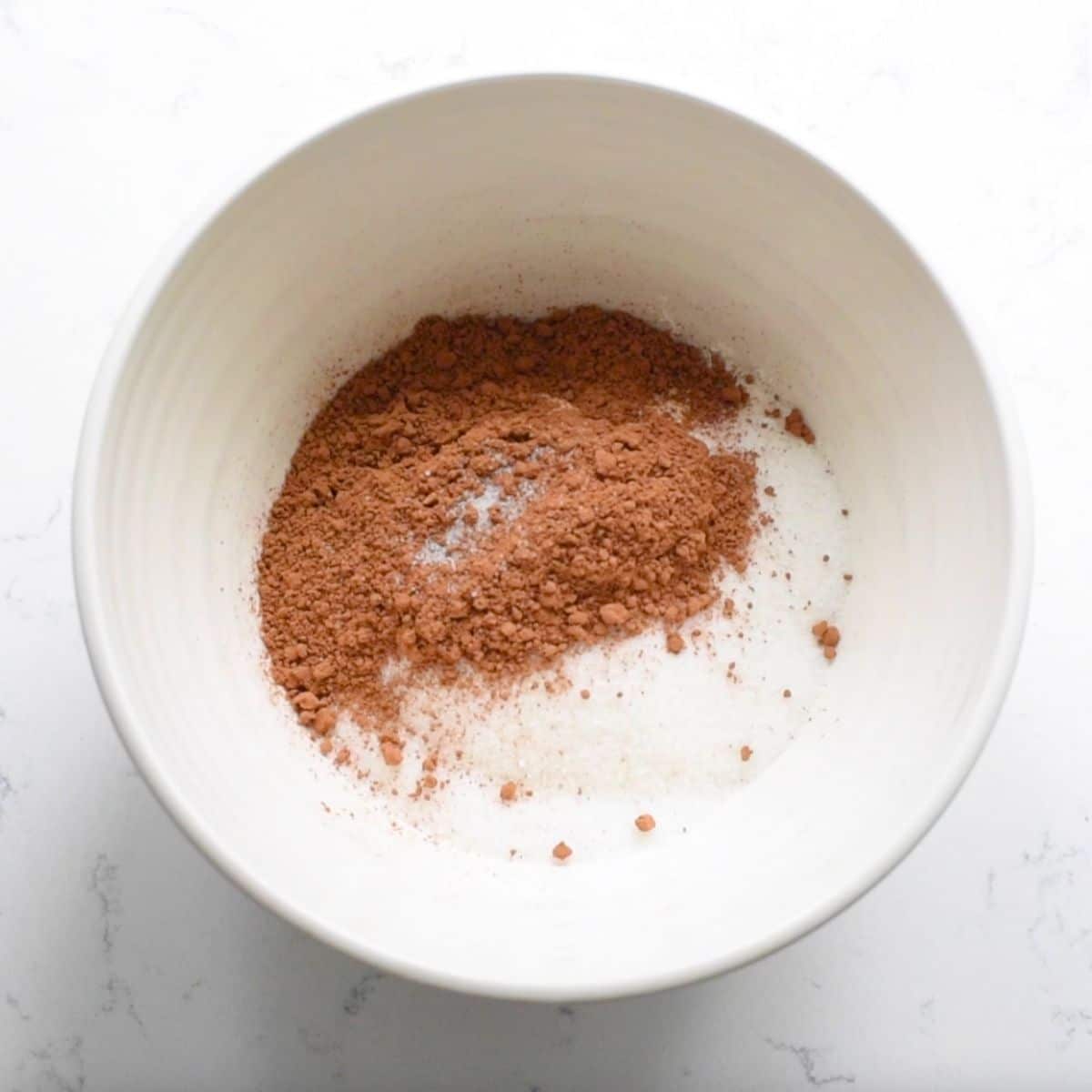 A white bowl on a marble surface contains a mix of dry ingredients for small batch brownies, including cocoa powder and sugar, all ready for baking preparation.