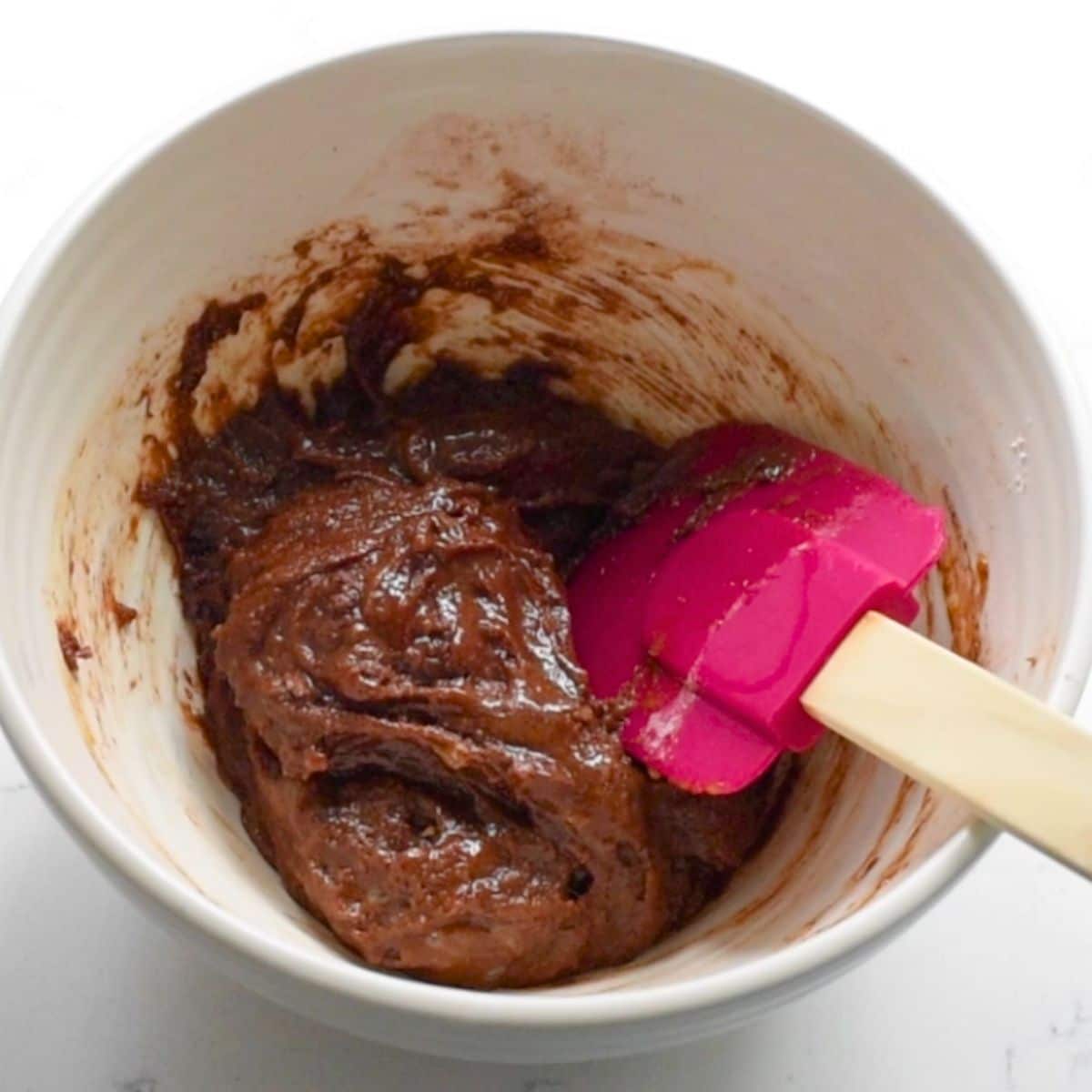 A white bowl holds a thick chocolate batter, perfect for small batch brownies. A pink spatula with a wooden handle rests inside, slightly smeared with batter. The scene is set against a simple white background.