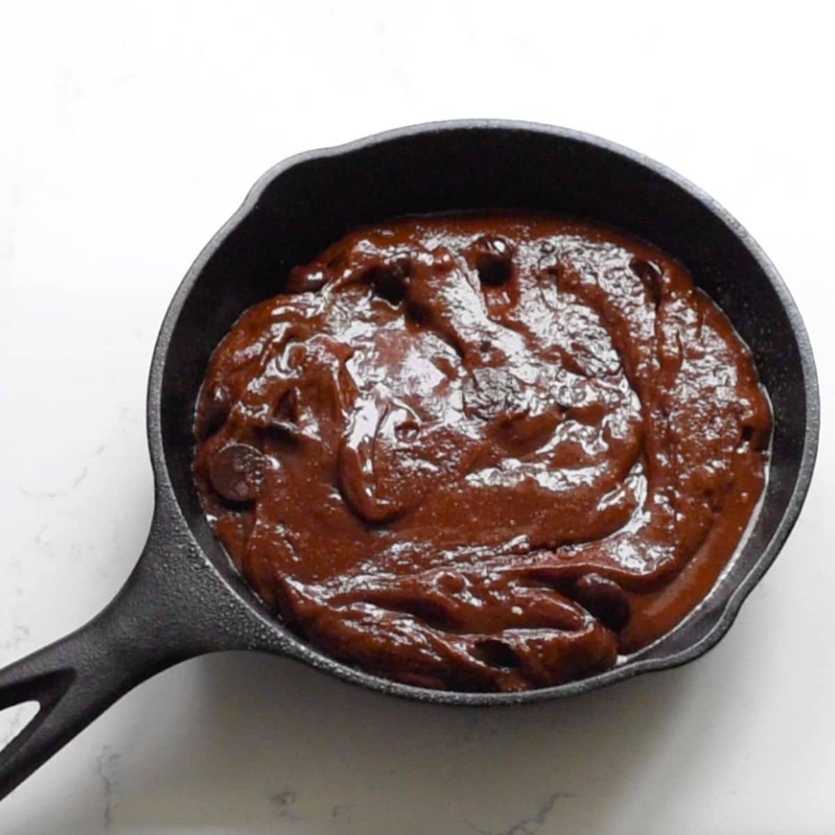 A cast iron skillet filled with unbaked small batch brownie batter, showcasing a rich, dark chocolate hue and studded with chocolate chips, placed on a pristine white surface.