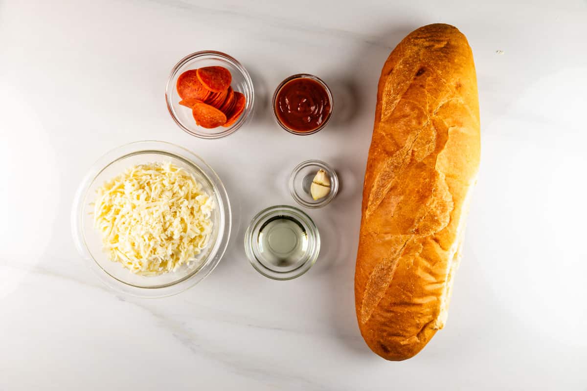 A loaf of French bread rests on a marble surface, surrounded by small bowls of sliced pepperoni, marinara sauce, shredded cheese, a clove of garlic, and olive oil—perfect components for creating a delicious French bread pizza.