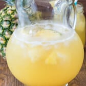 yellow party punch in a clear glass with pineapple in the back.