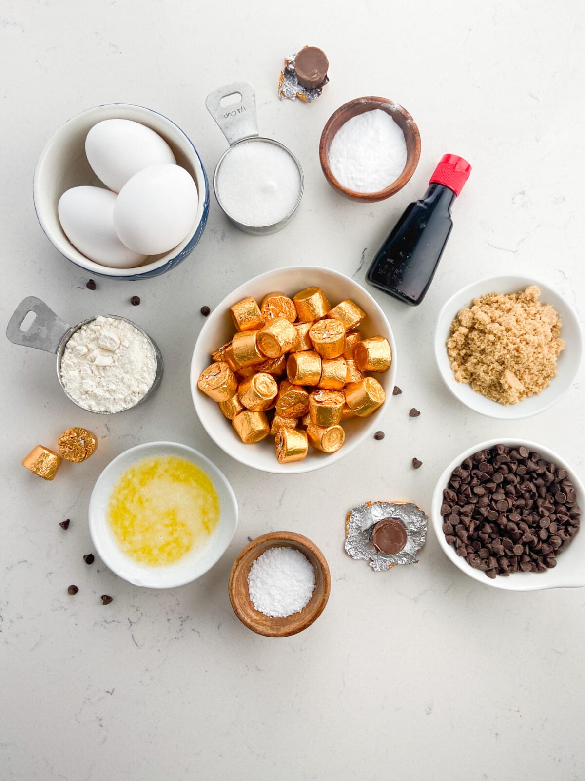 A flat lay of baking ingredients for Rolo cookies features eggs, sugar, baking soda, vanilla extract, flour, brown sugar, melted butter, salt, chocolate chips, and chocolate candies.