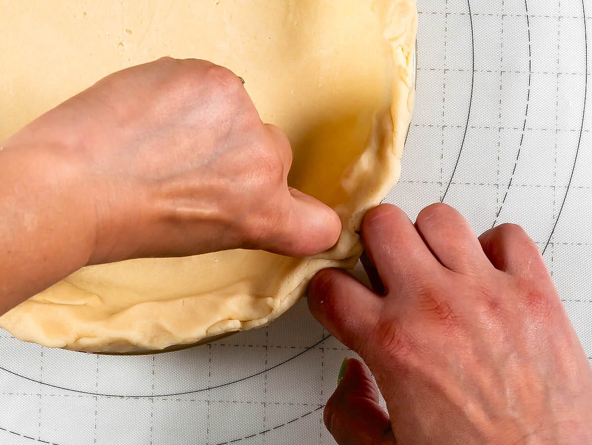 Close-up of hands crimping the edges of a pie crust on a dough mat with a circular design, following a classic pie crust recipe. The dough appears smooth and lightly golden, ready for baking.