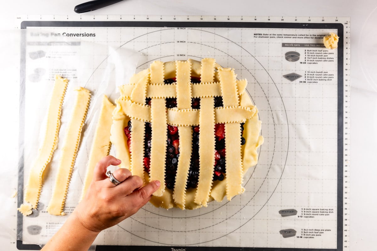 pie with fruit filling topped with vertical strips of pie dough and a few weaved horizontally with hand holding one strip.