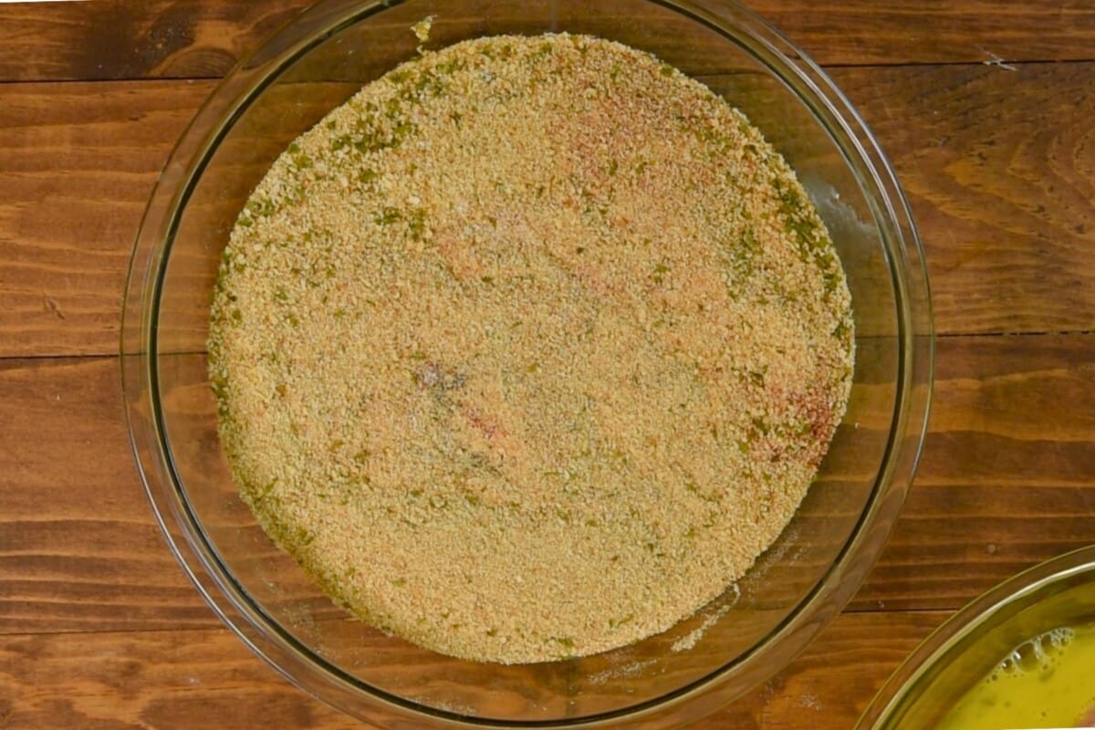 A glass bowl filled with seasoned breadcrumbs, perfect for oven-baked chicken, sits on a wooden surface. The breadcrumbs display a vibrant mixture of speckles and colors, hinting at the spices and herbs blended in.