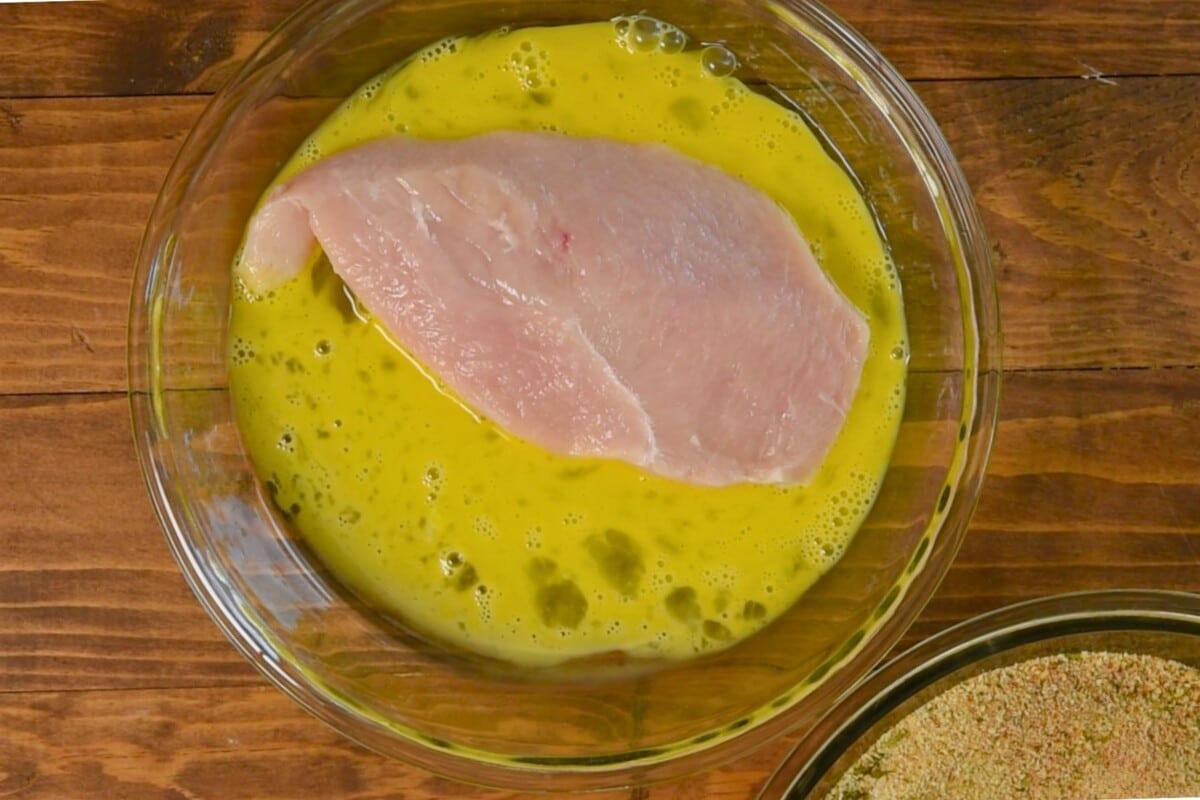 A raw chicken breast is submerged in a bowl of beaten eggs on a wooden surface, ready to become delicious oven baked chicken. A bowl of breadcrumbs is partially visible in the lower right corner.