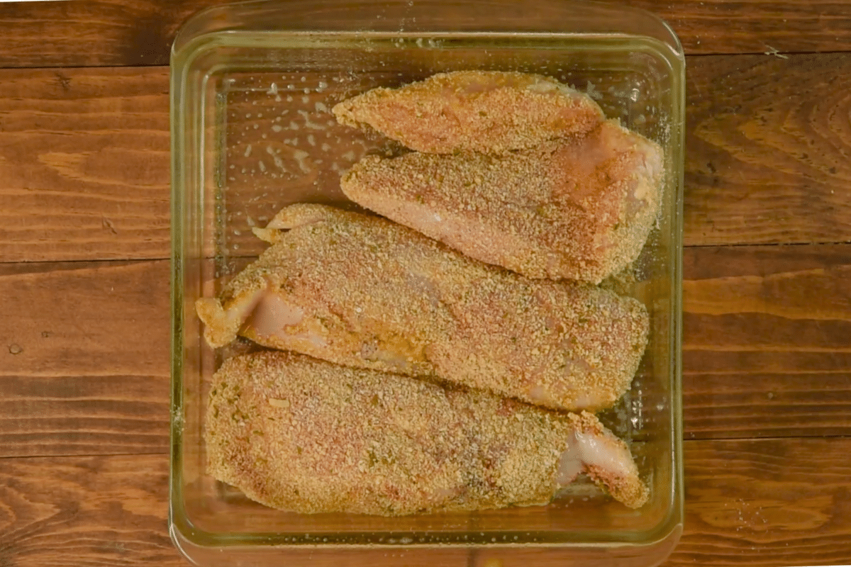 Four breaded chicken breasts are arranged in a glass baking dish on a wooden surface, ready to become delicious oven-baked chicken. Coated with a grainy, seasoned breadcrumb mixture, these tender pieces are prepped for cooking perfection.