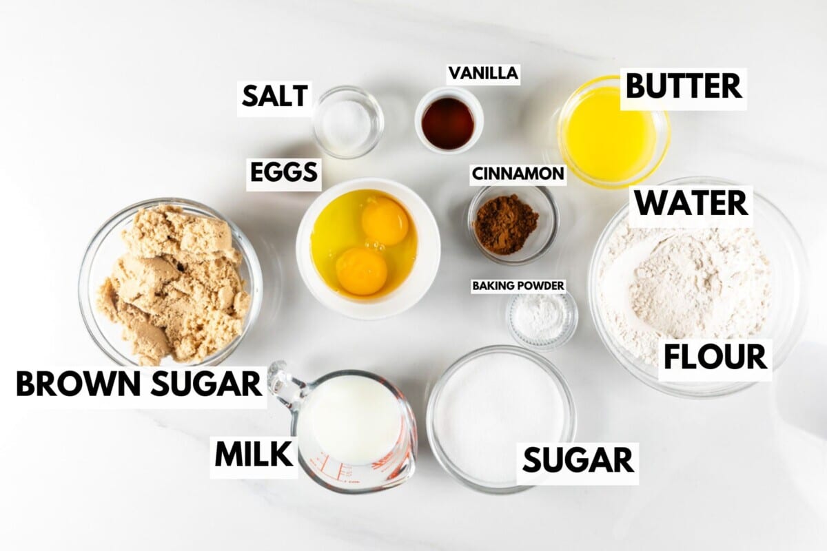 A collection of baking ingredients for a perfect coffee cake on a white surface, featuring bowls of brown sugar, flour, and sugar, a measuring cup of milk, eggs, small bowls of salt, vanilla, and cinnamon, melted butter, and water. Each item is meticulously labeled.