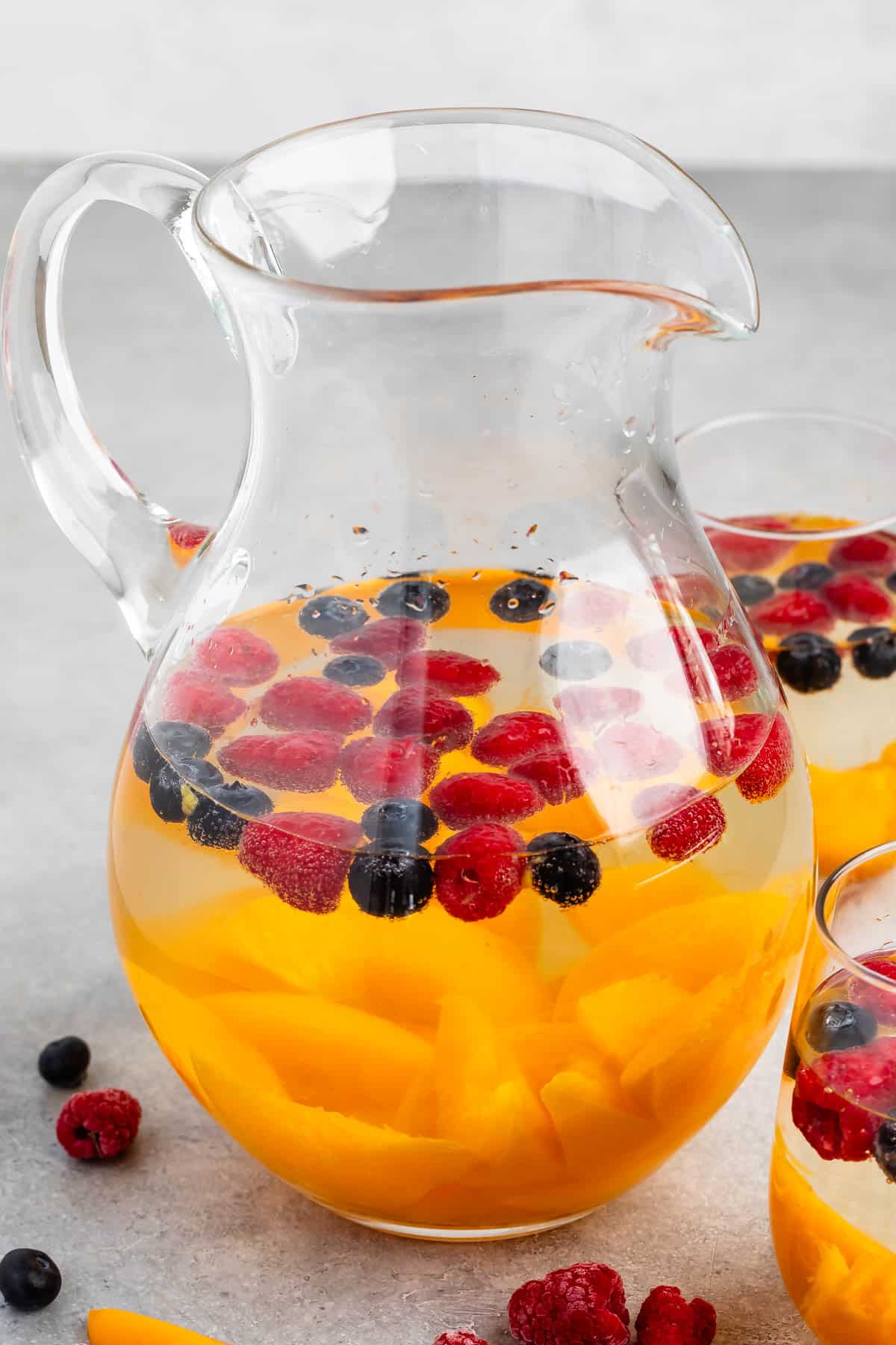 clear glasses holding the clear drink with raspberries, blueberries, and peaches sitting inside.