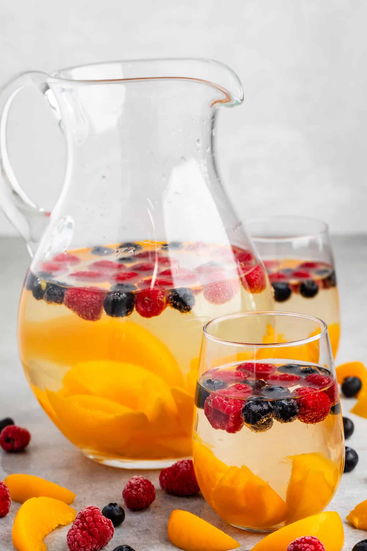 clear glasses holding the clear drink with raspberries, blueberries, and peaches sitting inside.