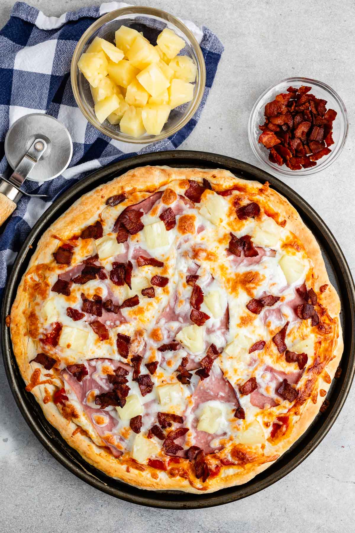 pizza with cheese, pineapple, ham, and bacon on top and bowls of pineapple and bacon above it.