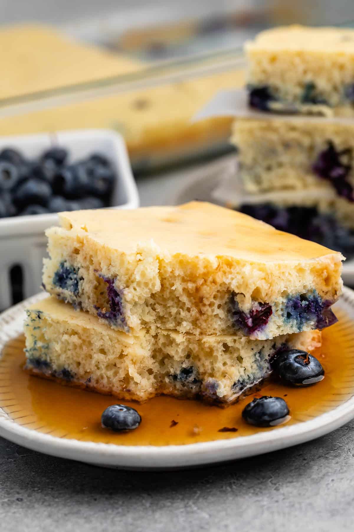 stacked square pancakes with blueberries baked in on a white plate covered in syrup.