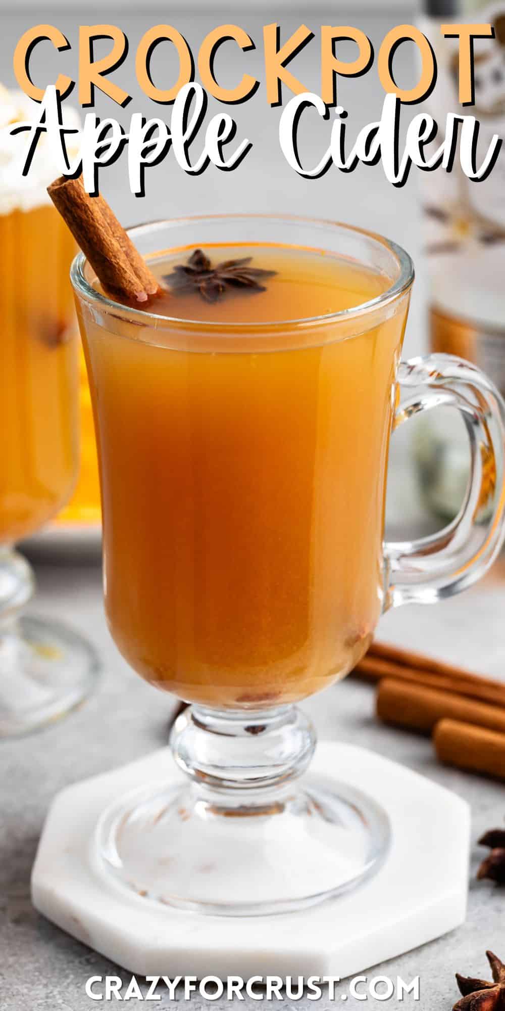 apple cider in a clear glass with a handle and a cinnamon stick and whipped cream on the top with words on the image.