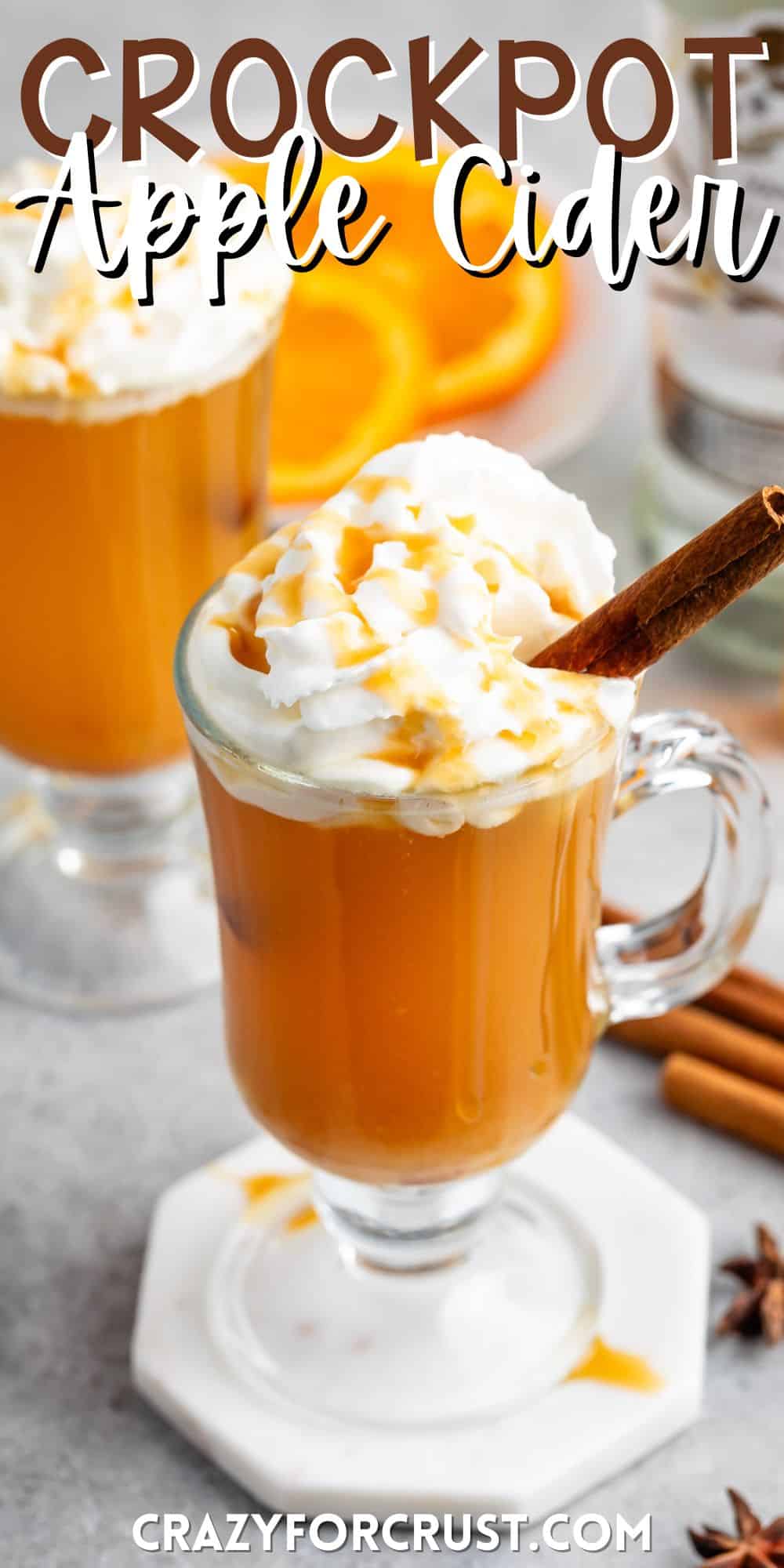 apple cider in a clear glass with a handle and a cinnamon stick and whipped cream on the top with words on the image.