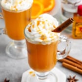 apple cider in a clear glass with a handle and a cinnamon stick and whipped cream on the top.