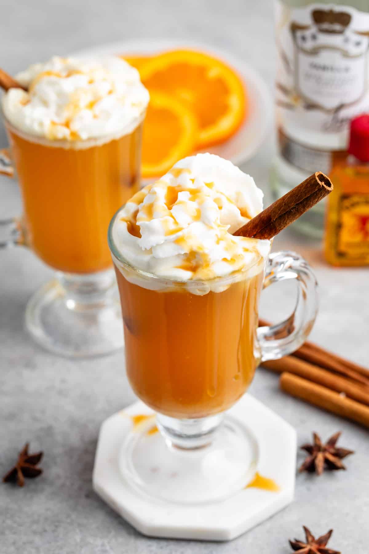 apple cider in a clear glass with a handle and a cinnamon stick and whipped cream on the top.