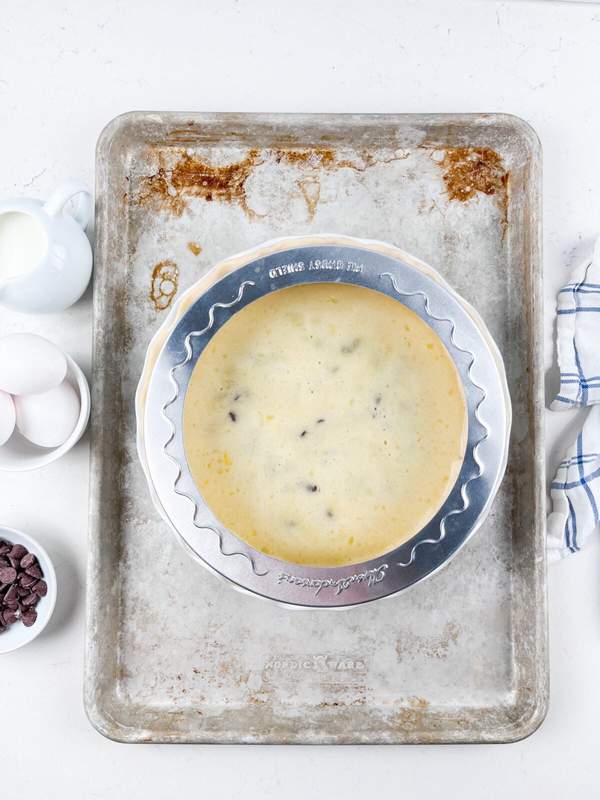 A pie crust filled with creamy batter and chocolate chips sits on a baking sheet. Surrounding items include a bowl of eggs, chocolate chips, a small pitcher of milk, and a white and blue checkered cloth.