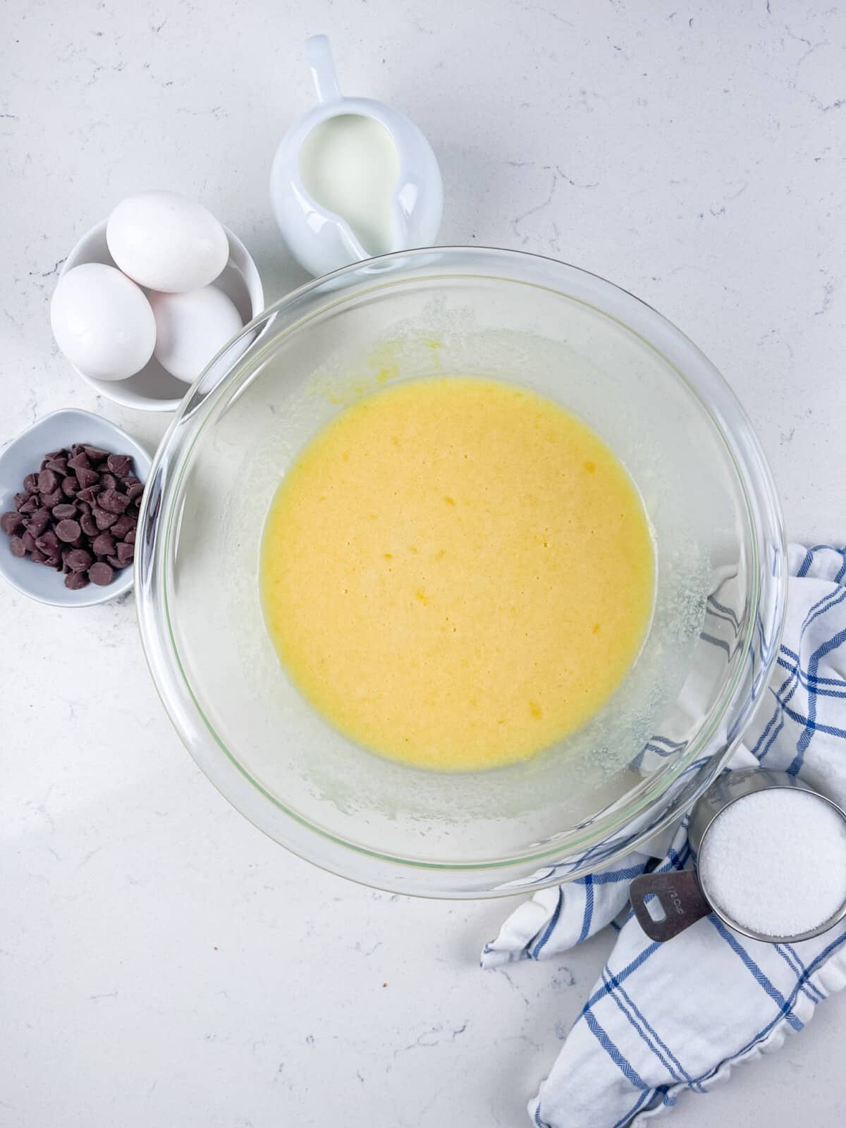 A glass bowl with a yellow batter mixture on a white surface, surrounded by a small pitcher of milk, three eggs, a bowl of chocolate chips, and a cup of sugar on a blue and white checkered cloth.